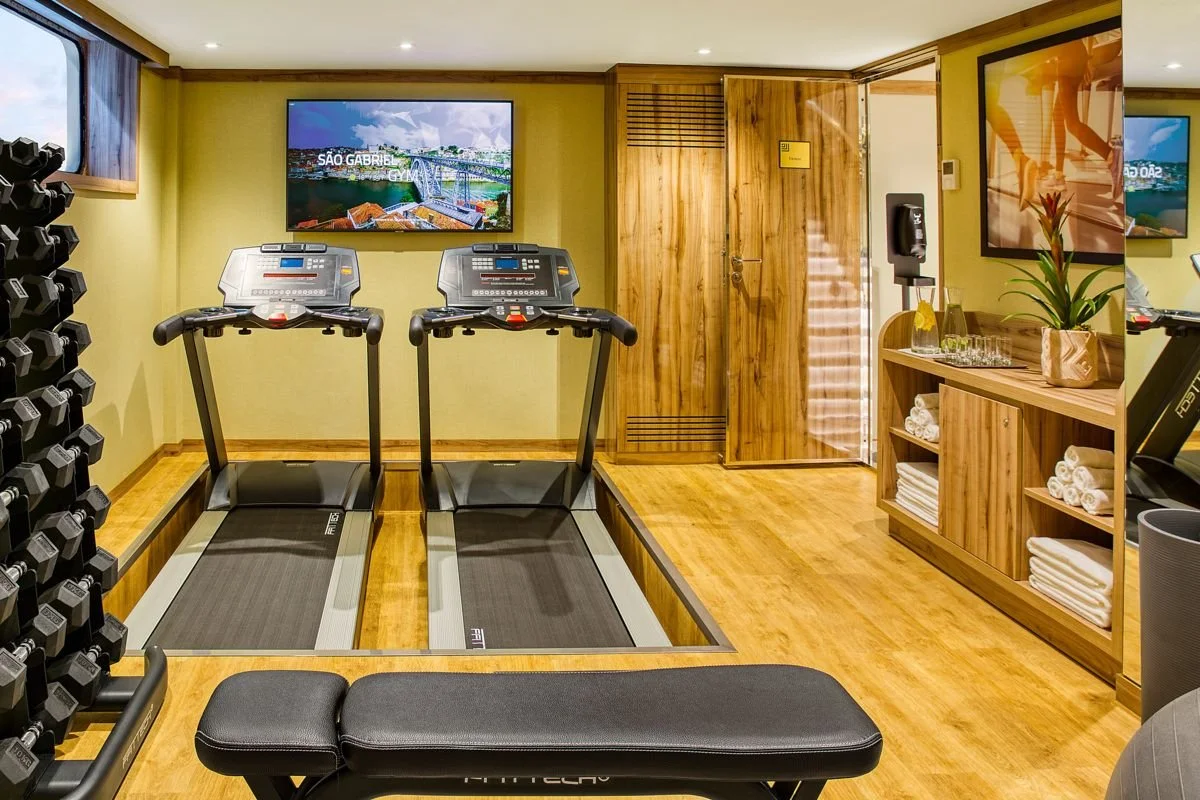 A small fitness room with two treadmills facing a wall-mounted TV, a rack of dumbbells to the left, a mirror on the right, and wooden shelves with rolled towels and decorative items. The room has wood flooring and soft lighting.