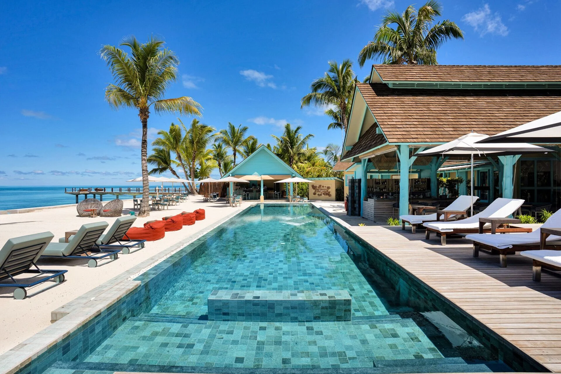 Beachside resort with a swimming pool surrounded by lounge chairs, umbrellas, palm trees, and resort buildings with a view of the ocean under a clear blue sky.