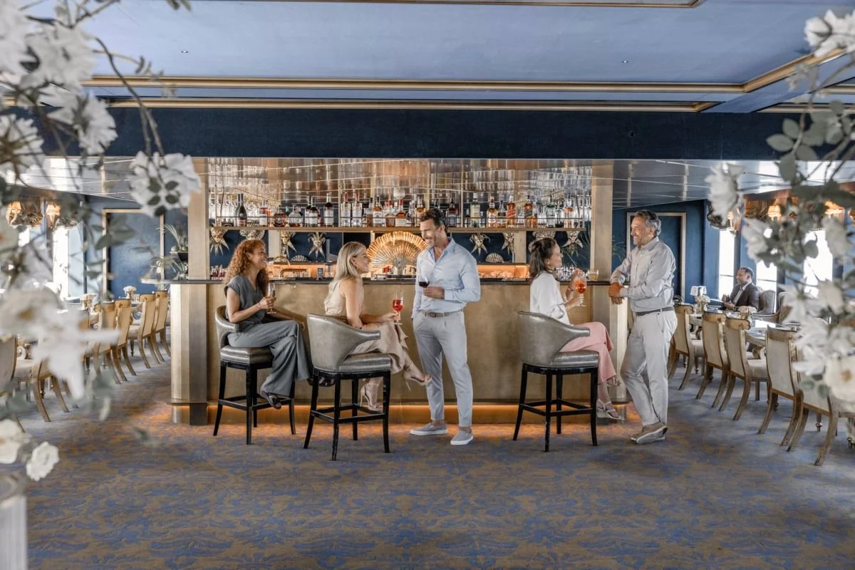A group of people socializing and drinking wine at a bar in a fancy restaurant with floral decorations and a blue and gold color scheme.