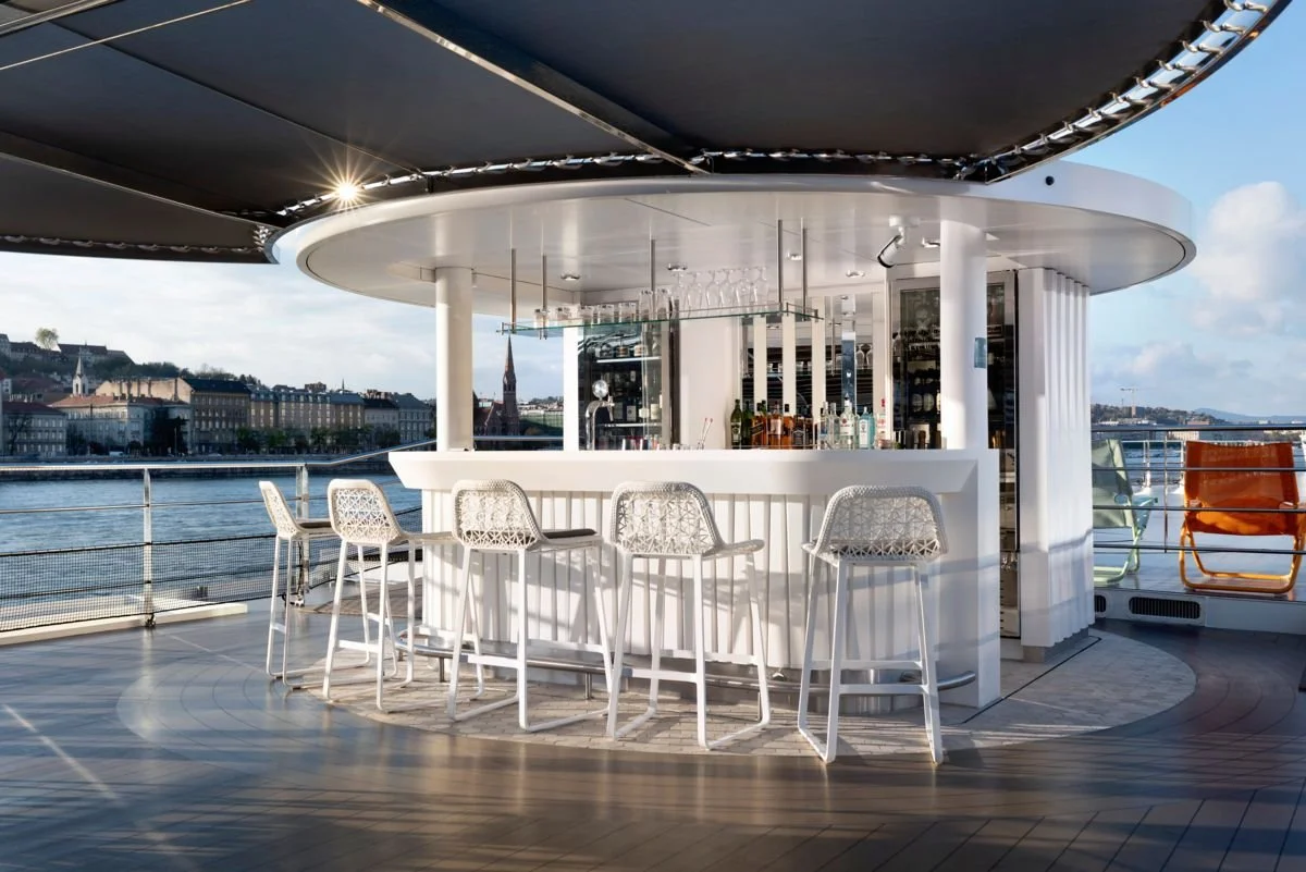 Outdoor bar area on a cruise ship deck with white stools and a canopy overhead, overlooking a river with buildings and a church steeple in the distance.