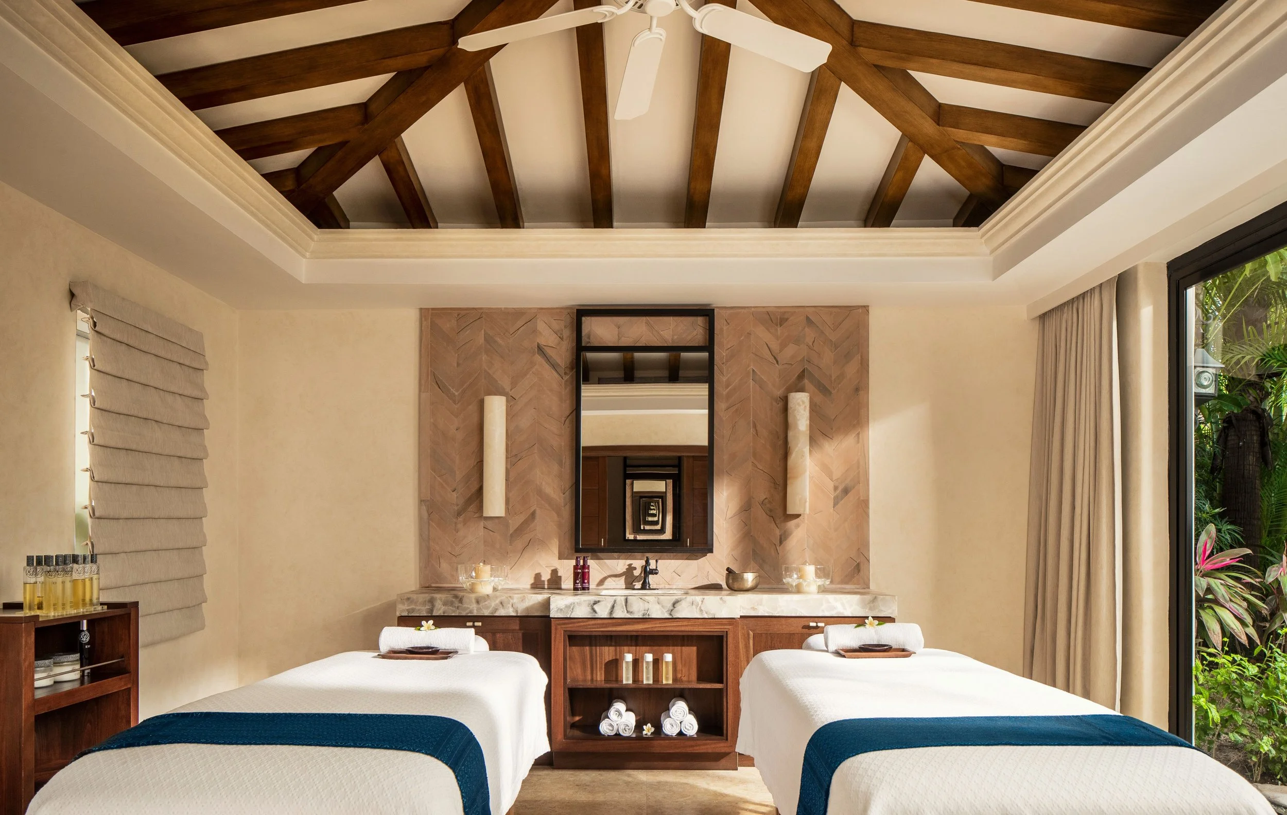 Spa room with two massage beds, a stone wall with a mirror and candles, and a ceiling fan in a room with beige walls, curtains, and natural light.