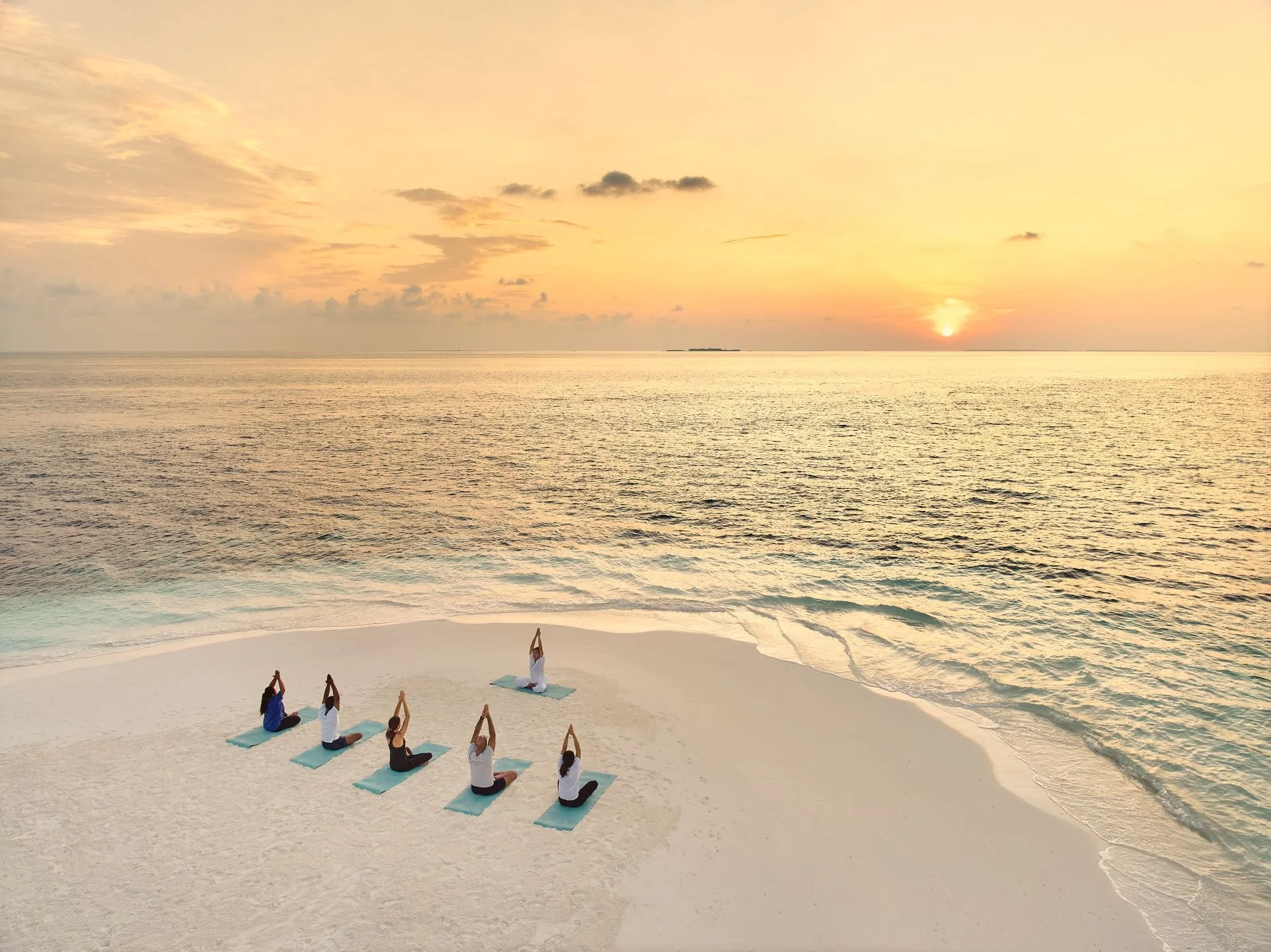 A group of people practicing yoga on a white sandy beach during sunset, with the ocean and a small island in the background.