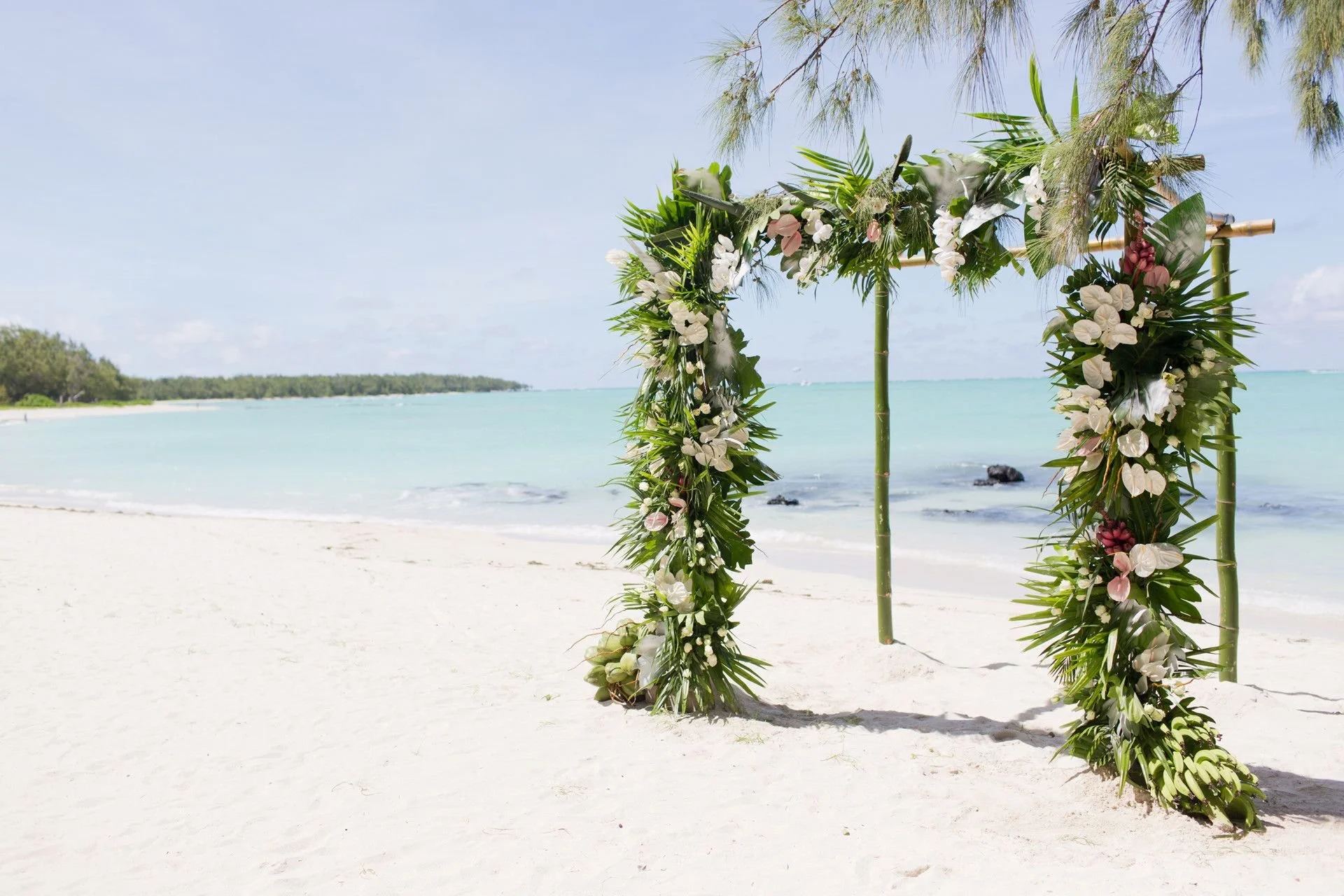 A decorative wedding arch made of bamboo and lush green foliage with white and pink flowers on a sandy beach by the ocean.
