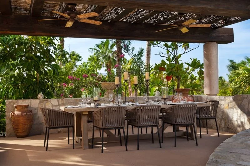 Outdoor dining area with a large wooden table, eight woven chairs, decorative candle holders, set with glassware and plates, surrounded by lush tropical plants and flowers, sheltered under a thatched roof.