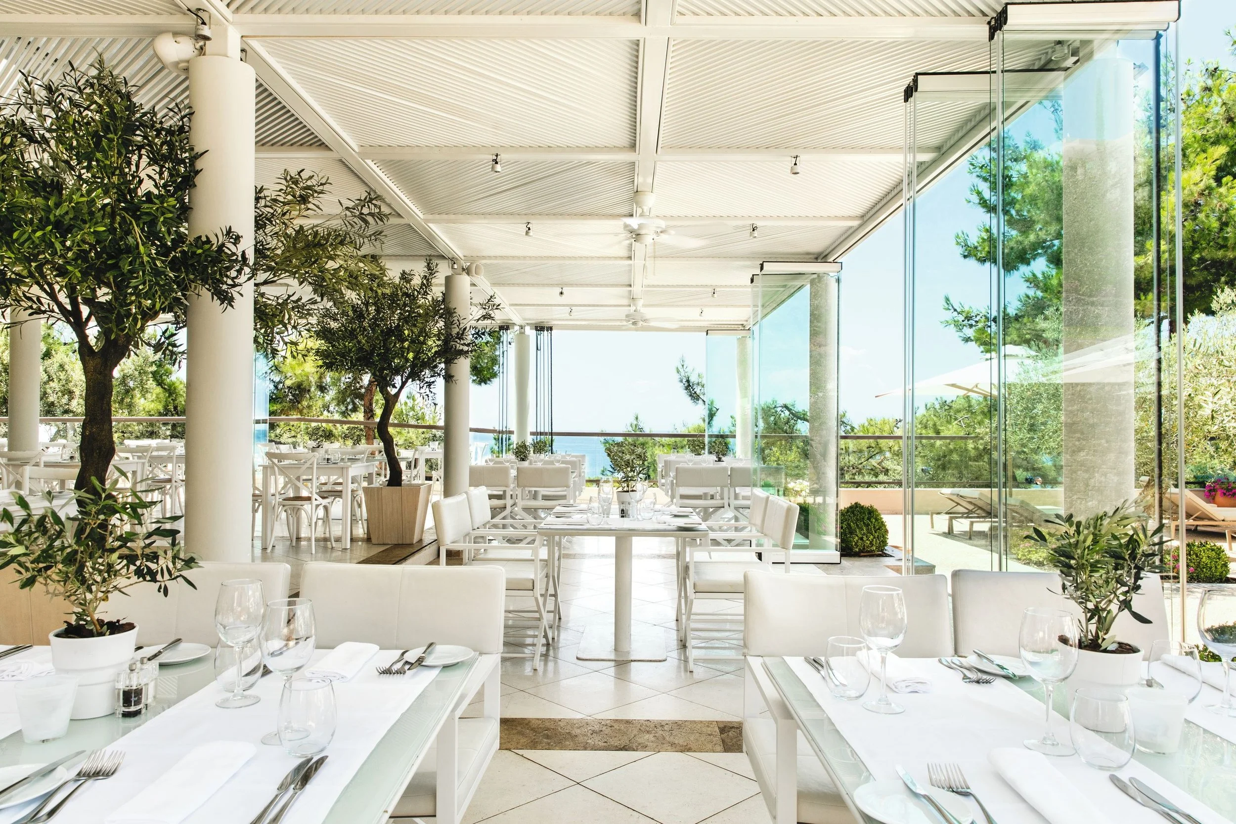Inside a bright, modern restaurant with white tables and chairs, potted plants, large glass windows, and a view of trees and the outdoors.