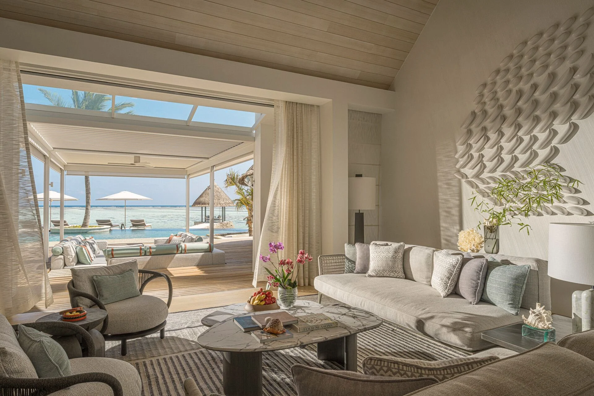 Living room with large open doors leading to a beach view with umbrellas and palm trees, decorated in neutral tones with modern furniture and indoor plants.