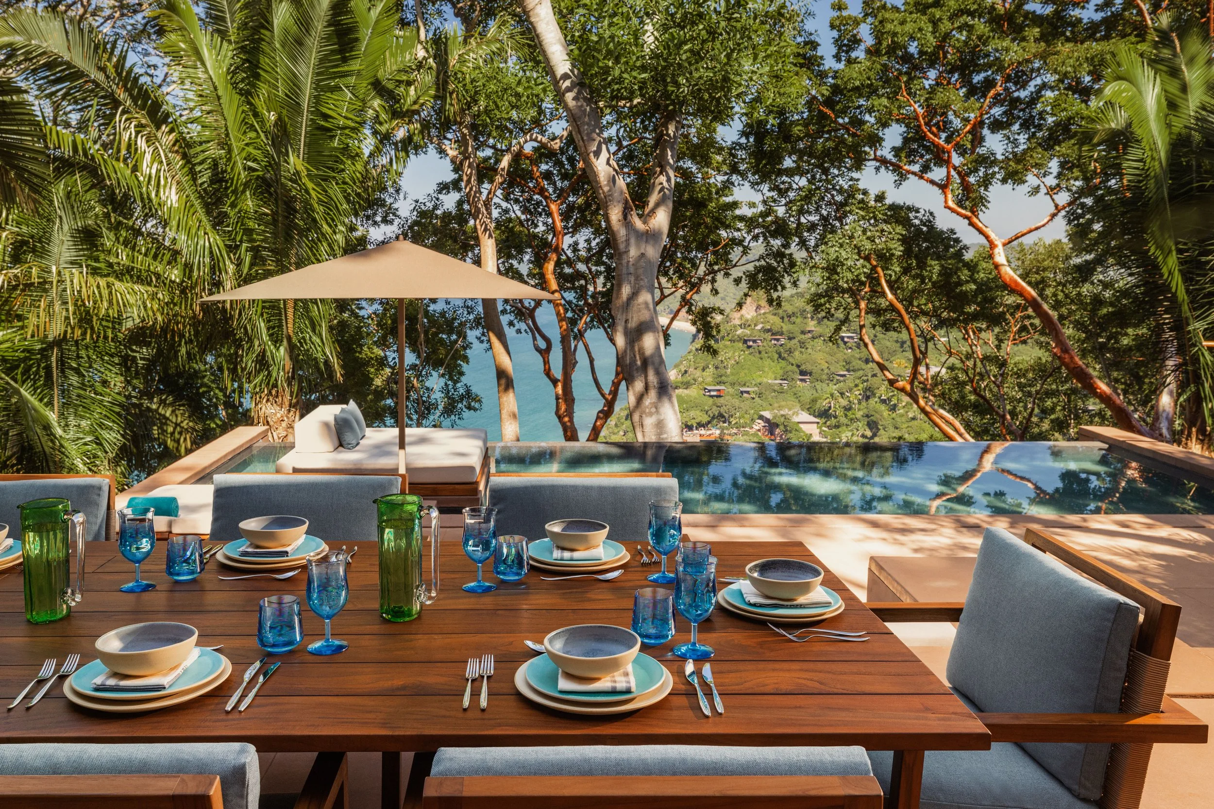 An outdoor dining table set with bowls, plates, glasses, and silverware, overlooking a tropical view with lush green trees, a large tree in the center, and a body of water in the background.