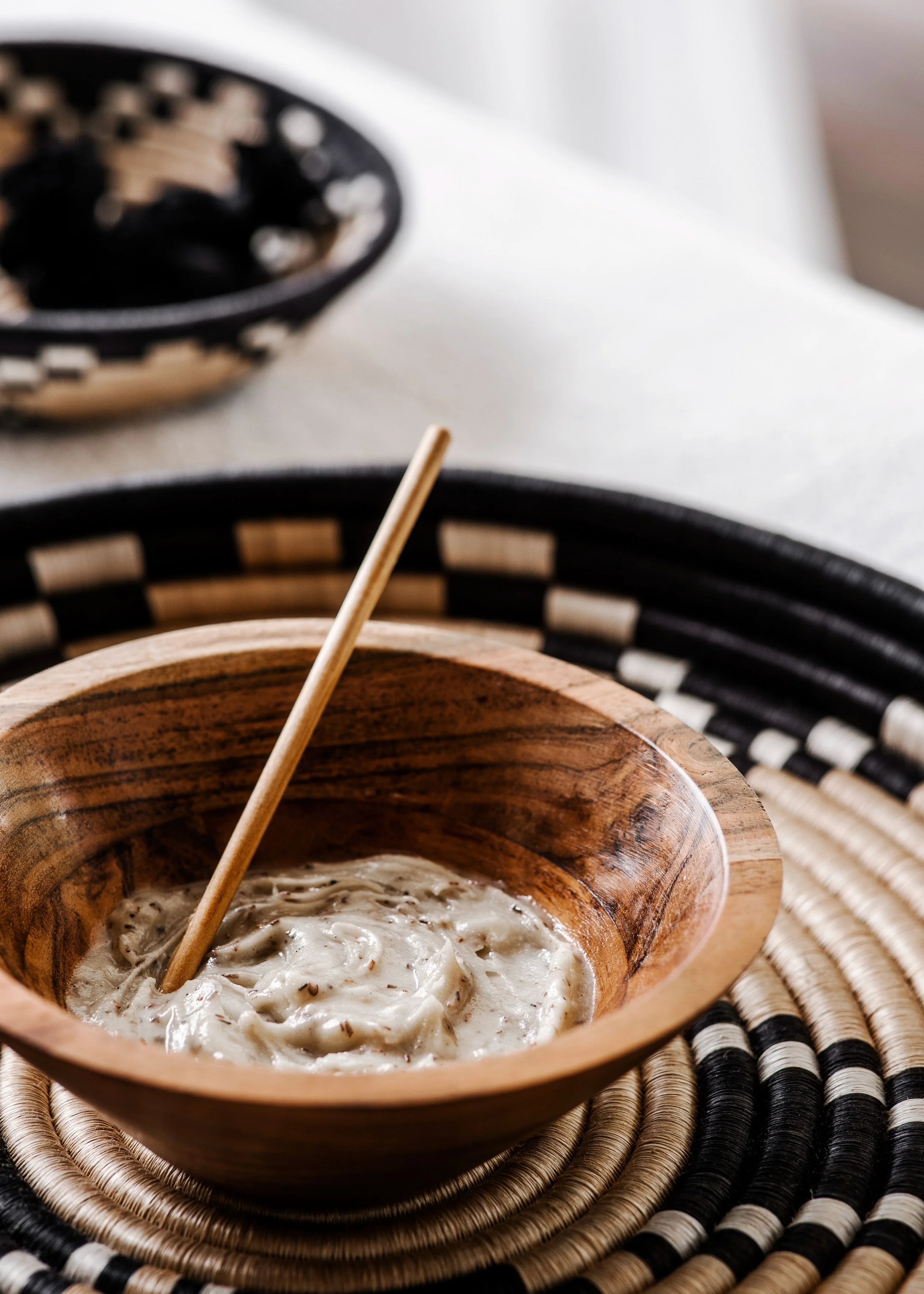 A wooden bowl containing a creamy, mushroom-based dish with a wooden skewer inside, placed on a black and natural woven placemat.