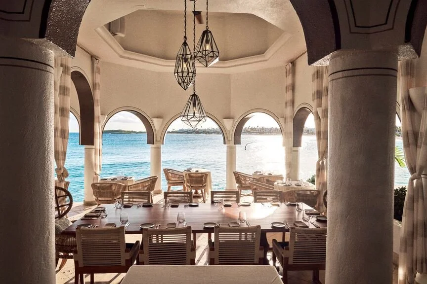 A restaurant with a long table set for dining, facing large arched windows that overlook a body of water, with wicker chairs and curtains, and decorative pendant lighting hanging from a ceiling.
