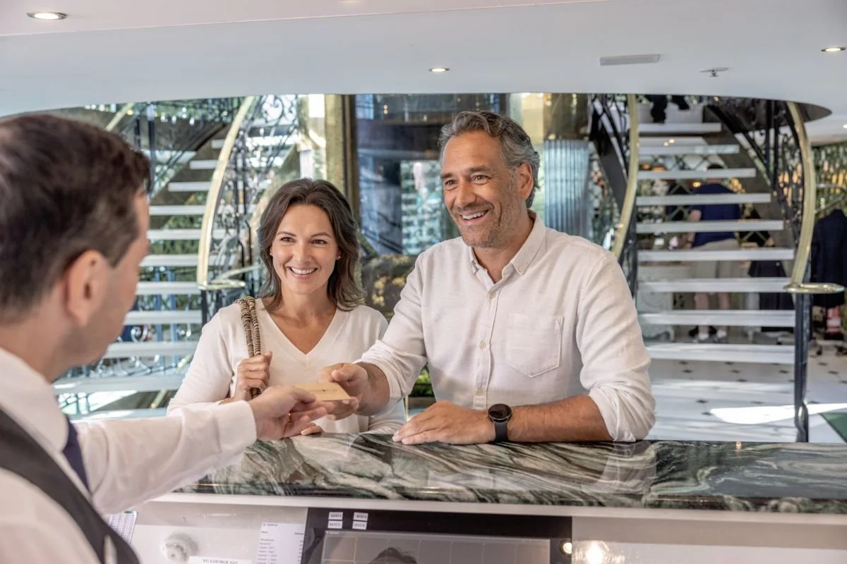 A man is checking into a hotel at the reception desk, handing over his credit card to the receptionist, while a couple smiles and waits.