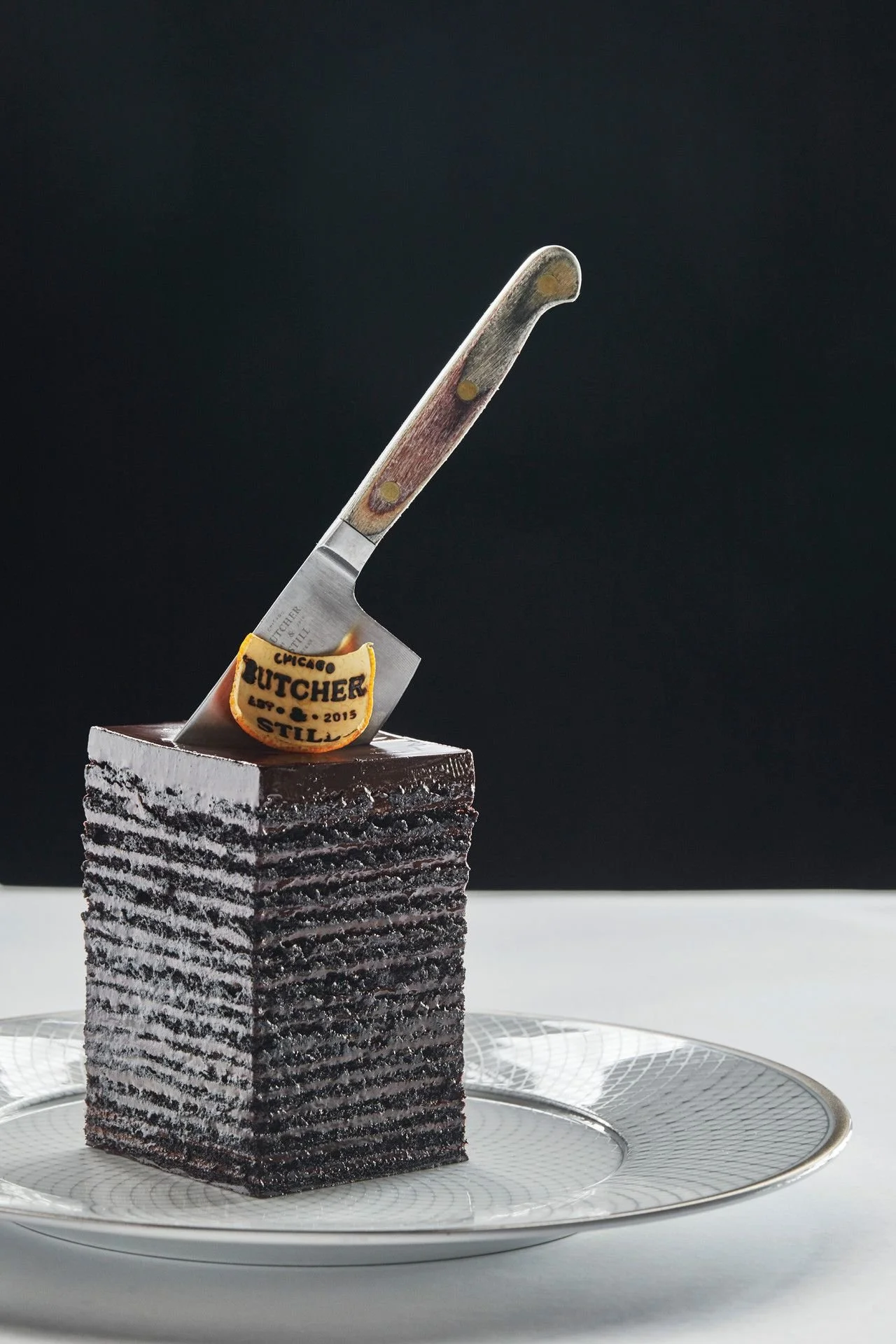 A multi-layered black and white layered cake with a small cutting knife inserted into the top. The knife has a wooden handle with a small leather patch that reads "Chicago Butcher Est. 2015". The cake is placed on a decorative silver tray.