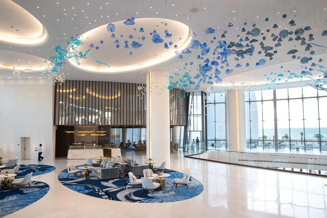 Interior of a modern hotel lobby with high ceilings, large windows overlooking the ocean, and blue glass sculptures hanging from the ceiling.