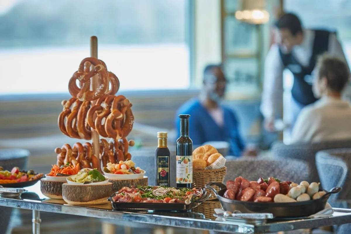 A table with various foods including pretzels on a wooden stand, salads, bread, sausages, and bottles of oil or vinegar, with people dining in the background.