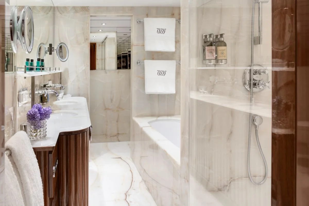 Luxurious bathroom with marble walls and floor, featuring a sink with a wooden cabinet, purple flowers in a glass vase, towel hooks, two white towels, a bathtub, and a shower area with silver fixtures and shampoo bottles.