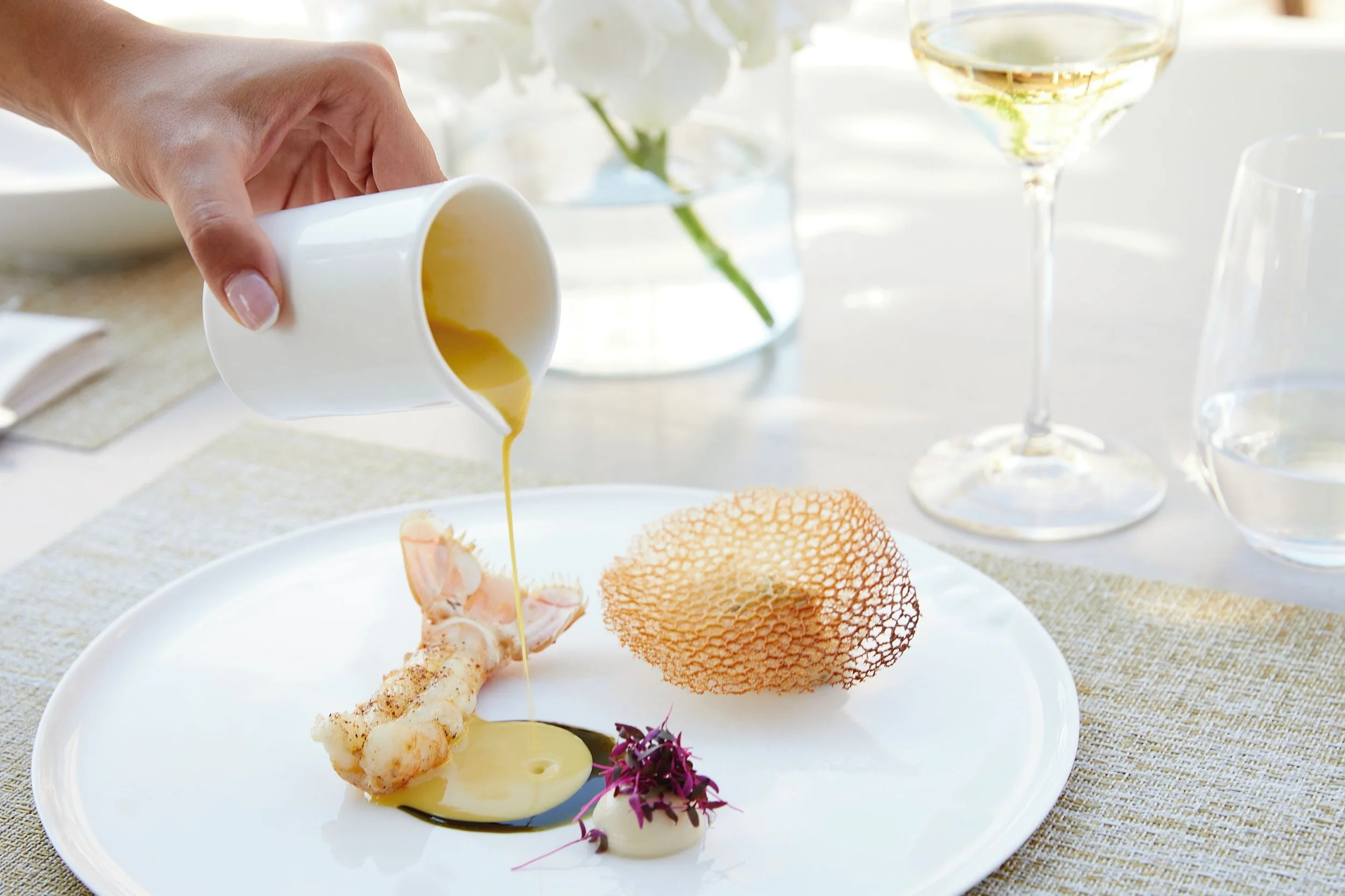 A person pouring yellow sauce over a gourmet seafood dish on a white plate with decorative elements, set on a table with glasses and flowers in the background.