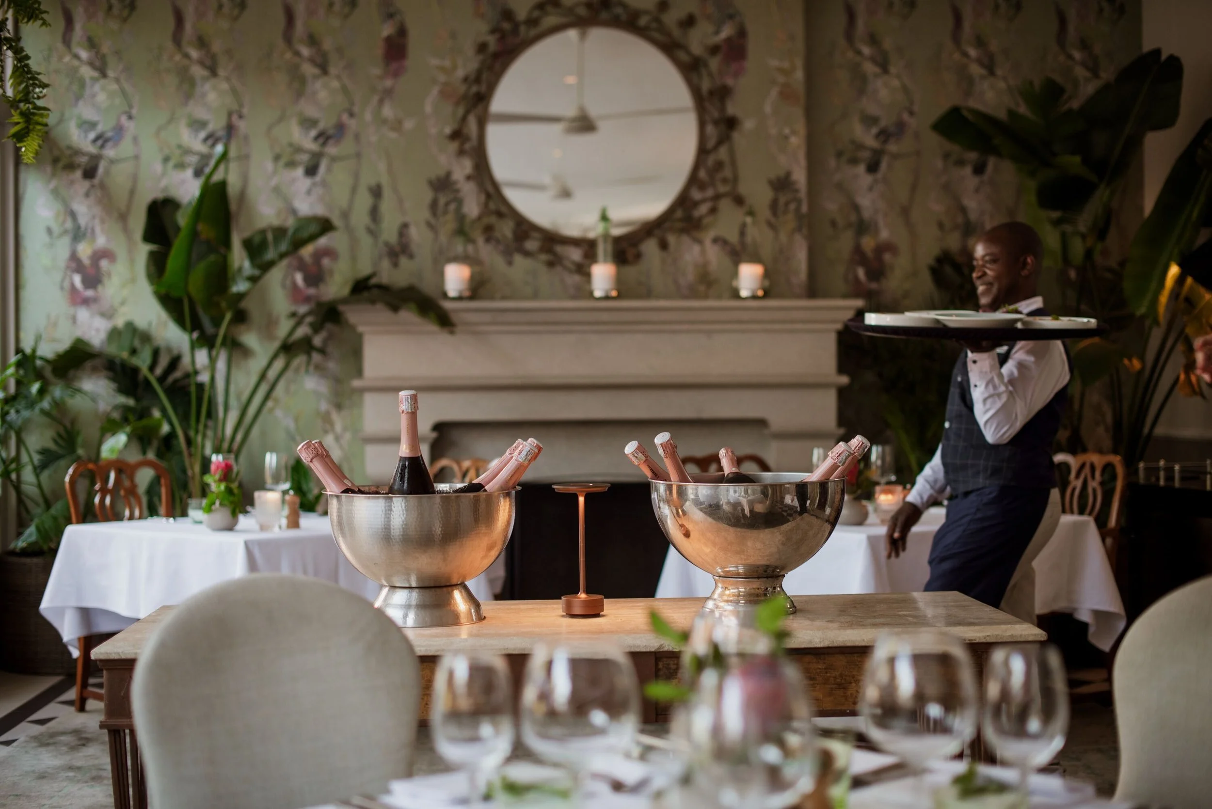 Elegant dining room with a large mirror, candles, potted plants, and tables set for a celebration, including bottles of champagne in coolers and glassware, with a smiling waiter in the background.