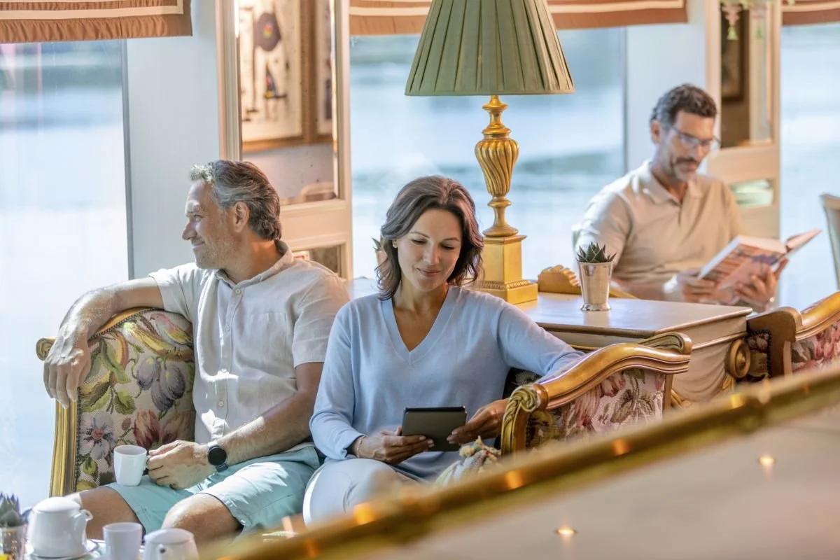 Three adults sitting on vintage furniture in a cozy, well-lit room, each engaged with different electronic devices, with a background of windows and decorative items.