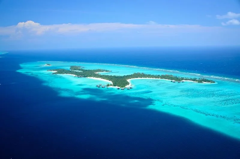 Aerial view of a tropical island with sandy beaches, lush green palm trees, and surrounding turquoise waters and coral reefs.