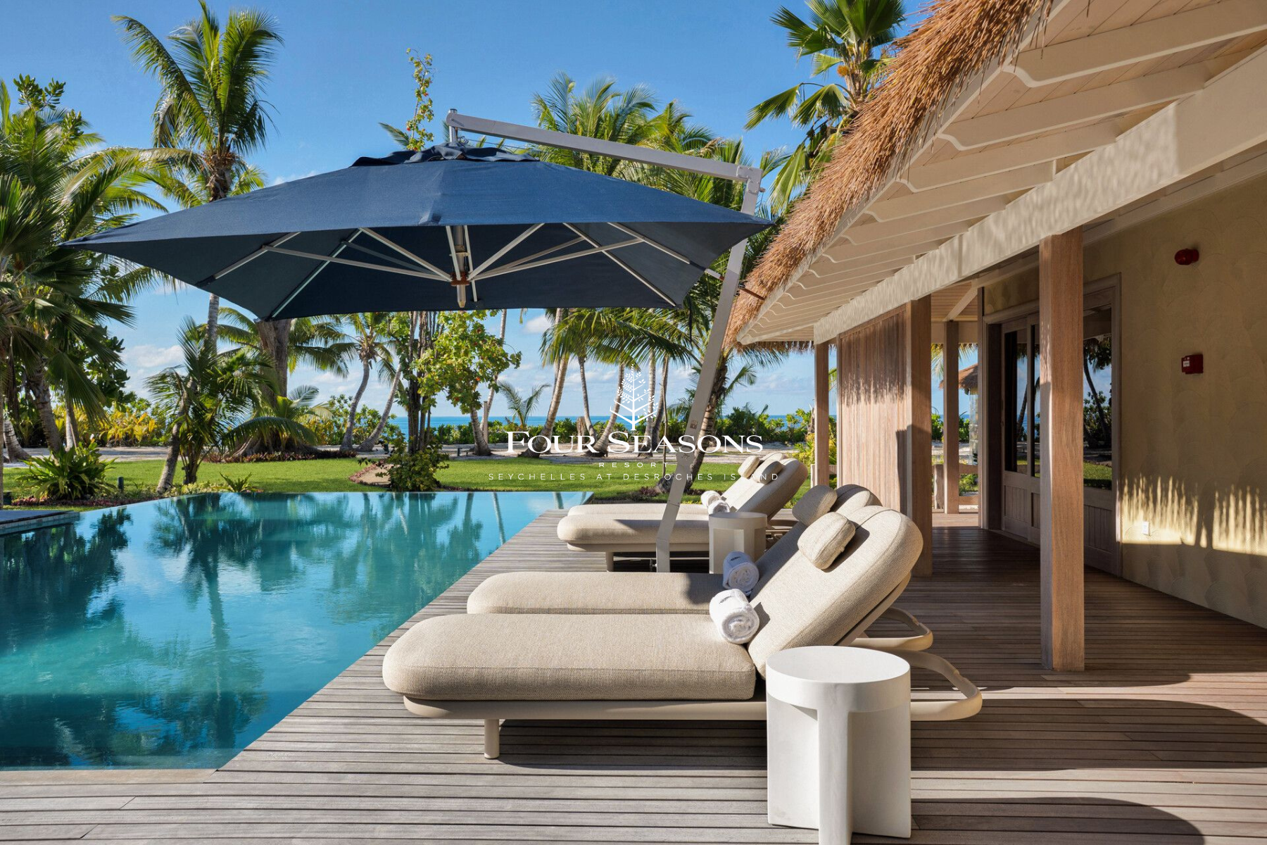 Poolside lounge chairs with rolled towels, sun umbrellas, and a thatched-roof building at a tropical resort with palm trees and ocean view in the background.