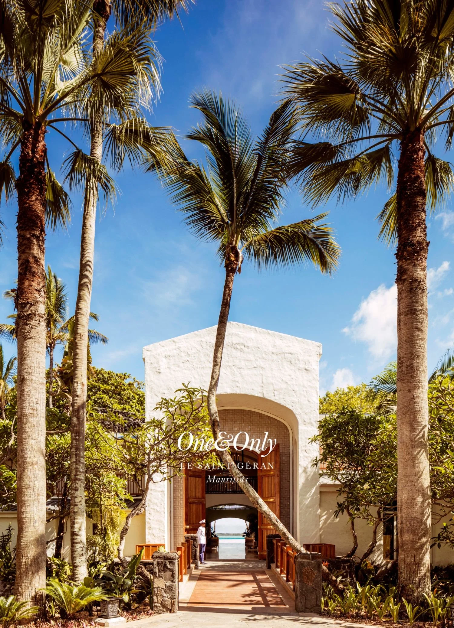 Entrance to a white building with an arched doorway framed by palm trees and lush greenery. The sky is clear and blue. Text on the image reads 'One & Only Le Saint Géran Mauritius.'