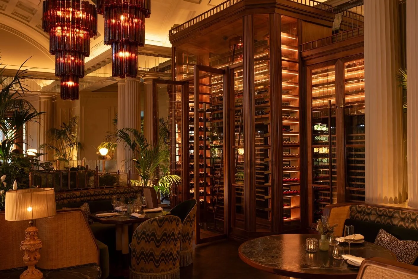 Elegant restaurant interior featuring a large wine storage area with glowing lighting, surrounded by lush green plants, tables with glassware and candles, plush chairs, and modern lamps, illuminated by warm lighting and a striking red ceiling chandelier.