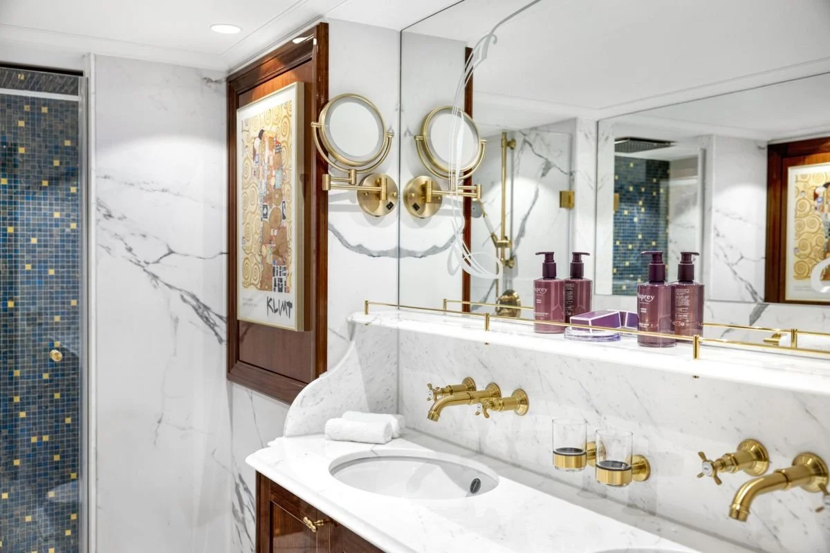 Luxurious bathroom with white marble walls and gold fixtures, including mirrors, faucets, and dispensers, decorated with purple toiletries and an abstract art piece on the wall.
