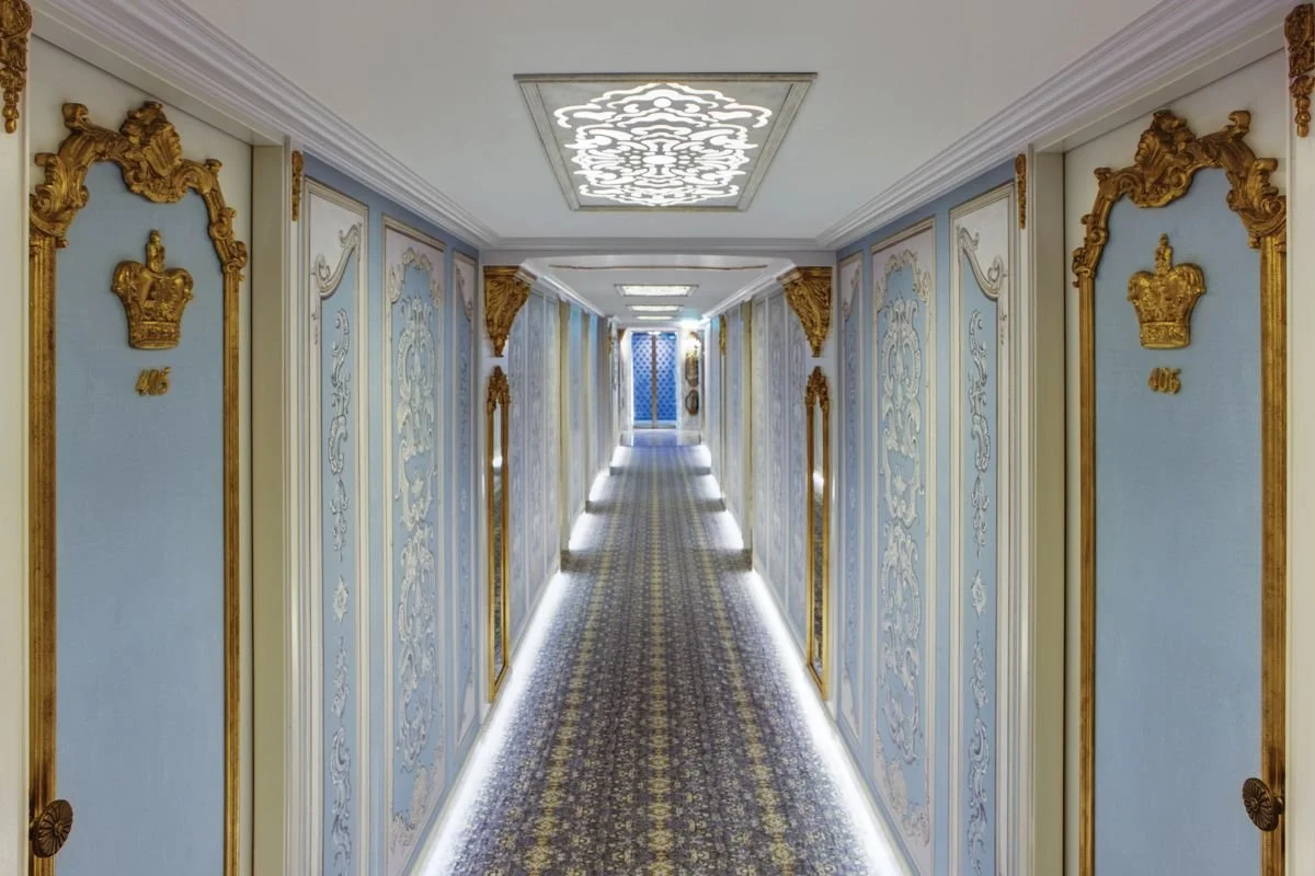 Elegant hotel corridor with ornate gold decorative accents on light blue and white walls, and a patterned carpet. The corridor is illuminated by ceiling lights and LED strip lighting along the base of the walls.