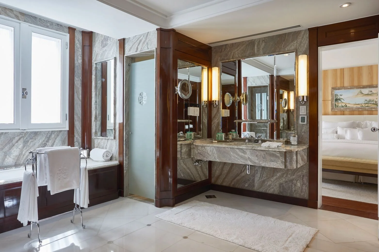 Luxury bathroom with marble countertops, a large mirror, and a towel rack with white towels. There is a frosted glass door and a window letting in natural light, with a view into a bedroom with a bed and artwork on the wall.