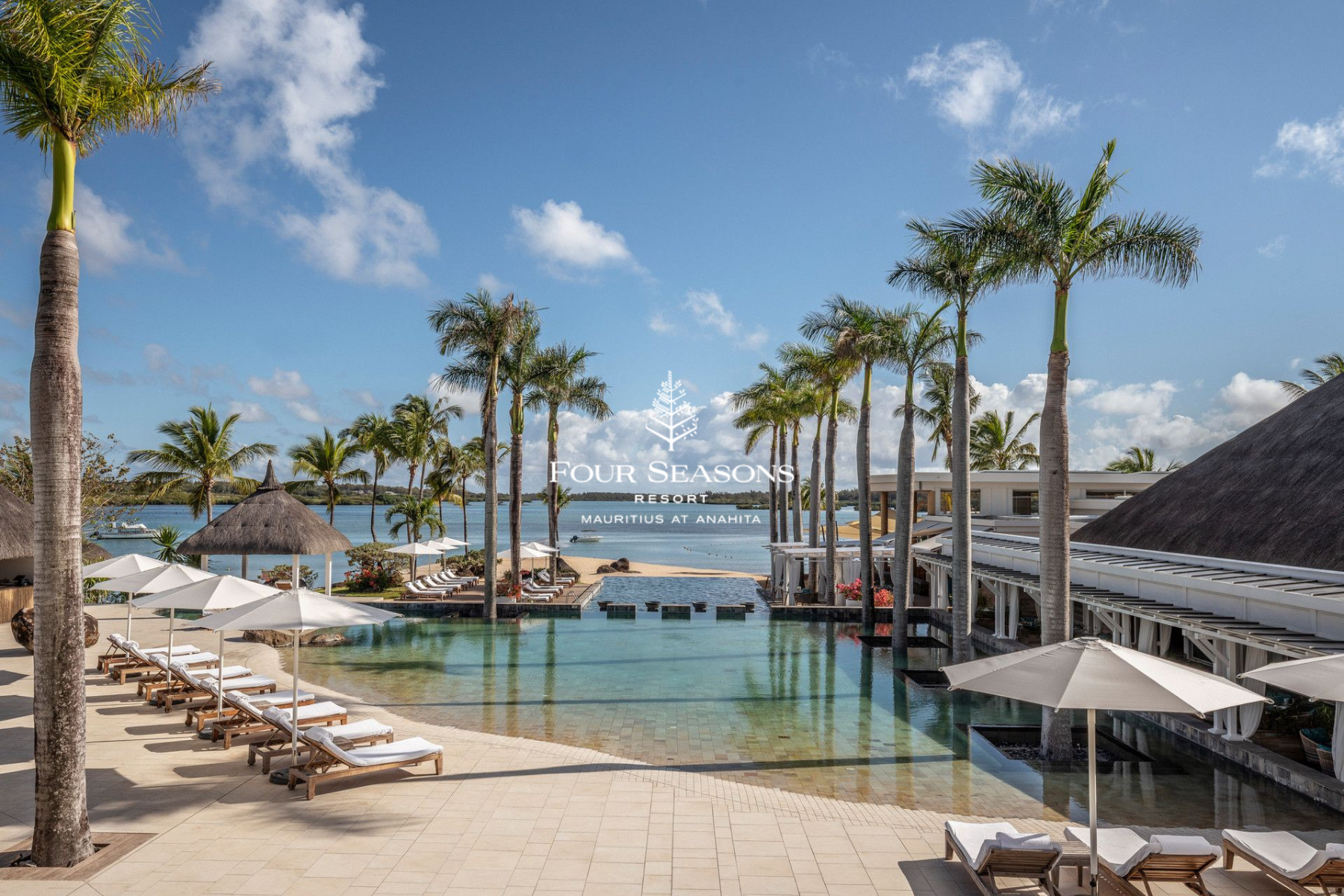 Resort pool area with lounge chairs under umbrellas, tall palm trees, thatched roof structures, and a view of the water with boats, under a partly cloudy blue sky. Overlaid text reads 'Four Seasons Resort Mauritius at Anahita'.