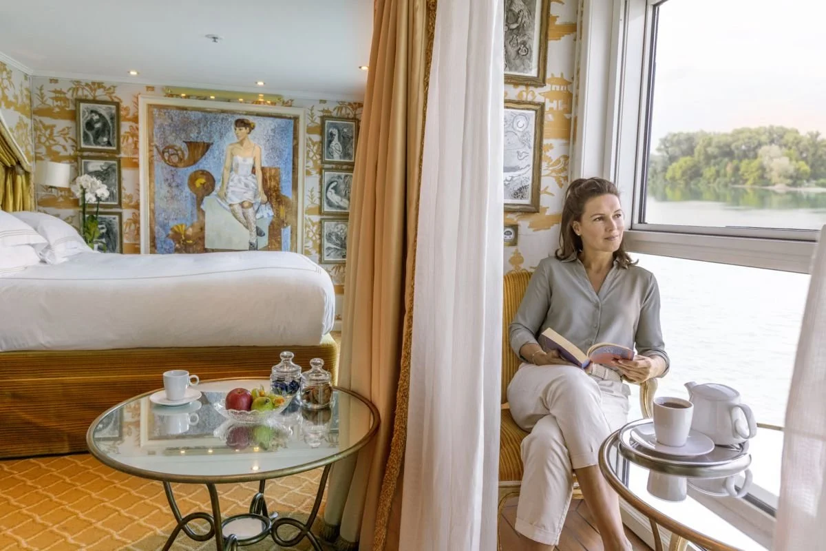 A woman sitting in a hotel room by the window, holding a book, with a tray of coffee and a teapot on a side table. The hotel room has a large bed with white linens, framed artwork on the wall, and a banana and apple on a glass-topped coffee table.