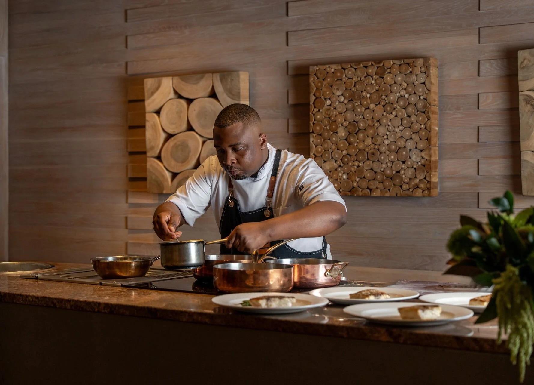 A male chef in a kitchen, wearing a white chef's coat and black apron, tending to food on the stove. The background features wooden wall art with logs, and a countertop with plates of prepared dishes and a bouquet of flowers.