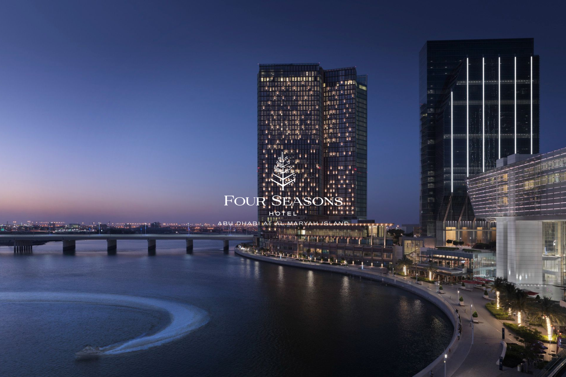 Night view of the Four Seasons hotel in Abu Dhabi at Al Maryah Island, featuring tall modern buildings illuminated with lights along a waterfront with a bridge and city lights in the background.