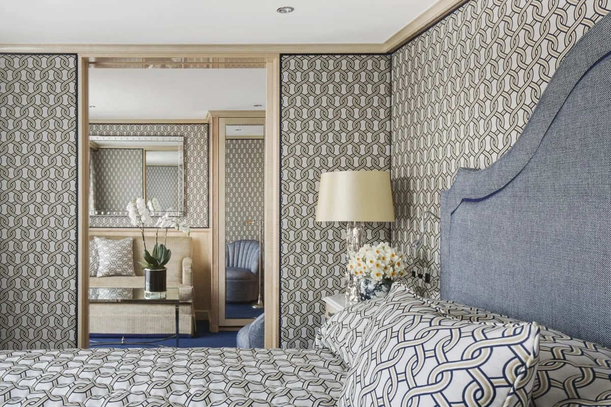 Hotel room interior with patterned wallpaper, a bed with a geometric-patterned comforter, a nightstand with a lamp and flowers, a mirror, a couch, and a mirror with a reflection of another room.