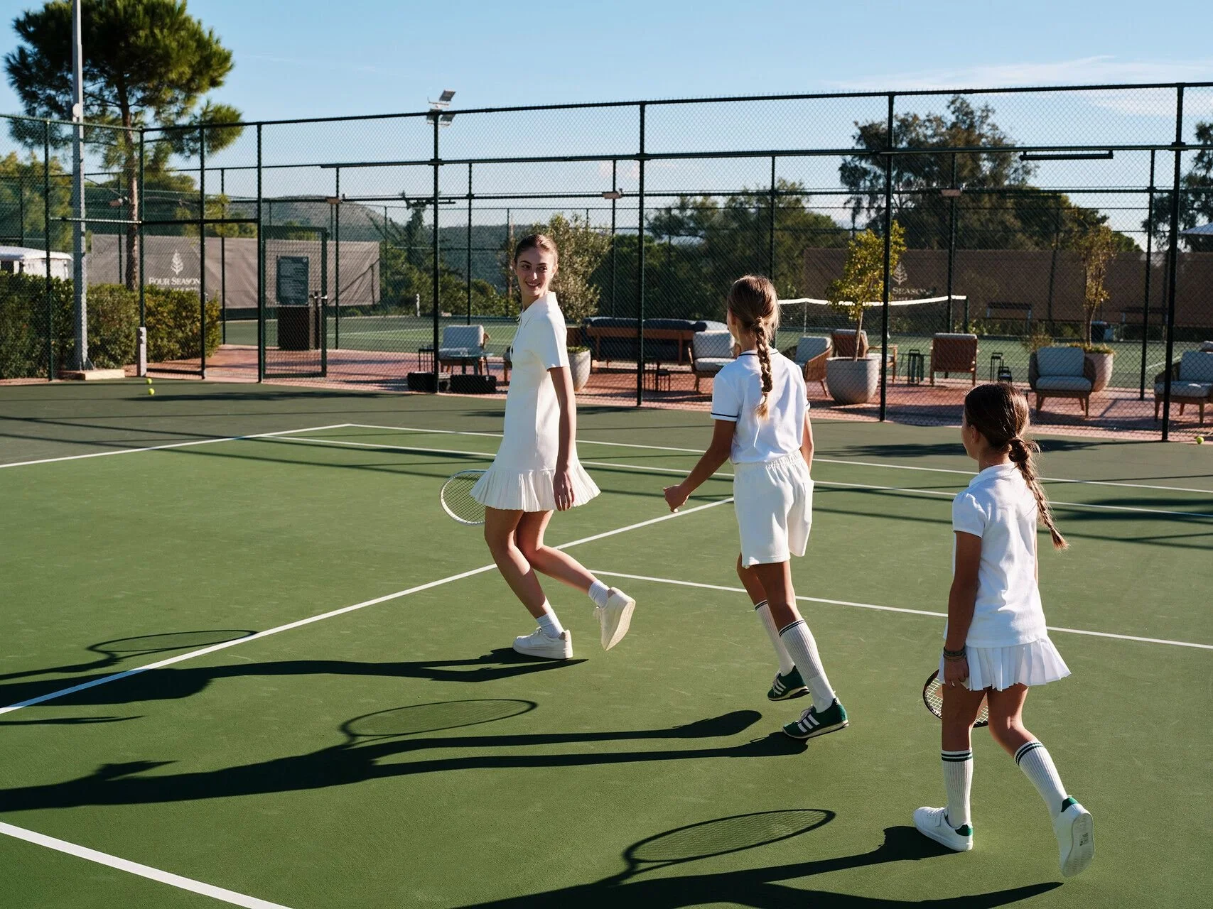 Three young girls in white tennis outfits on a tennis court, with one girl holding a tennis racket. They are outdoors on a sunny day with trees and seating in the background.