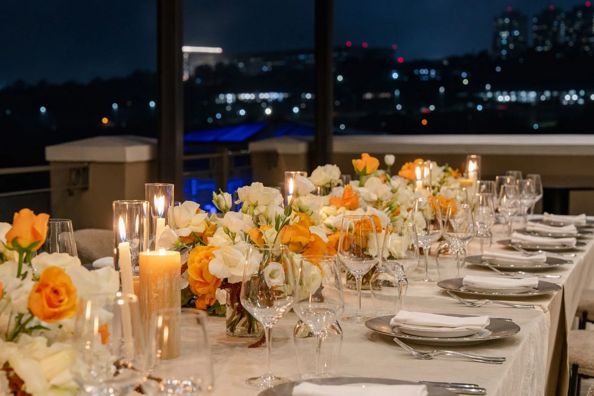 An elegantly set dinner table on a rooftop patio at night, decorated with white and orange flowers, candles, wine glasses, and place settings, with a city skyline illuminated in the background.