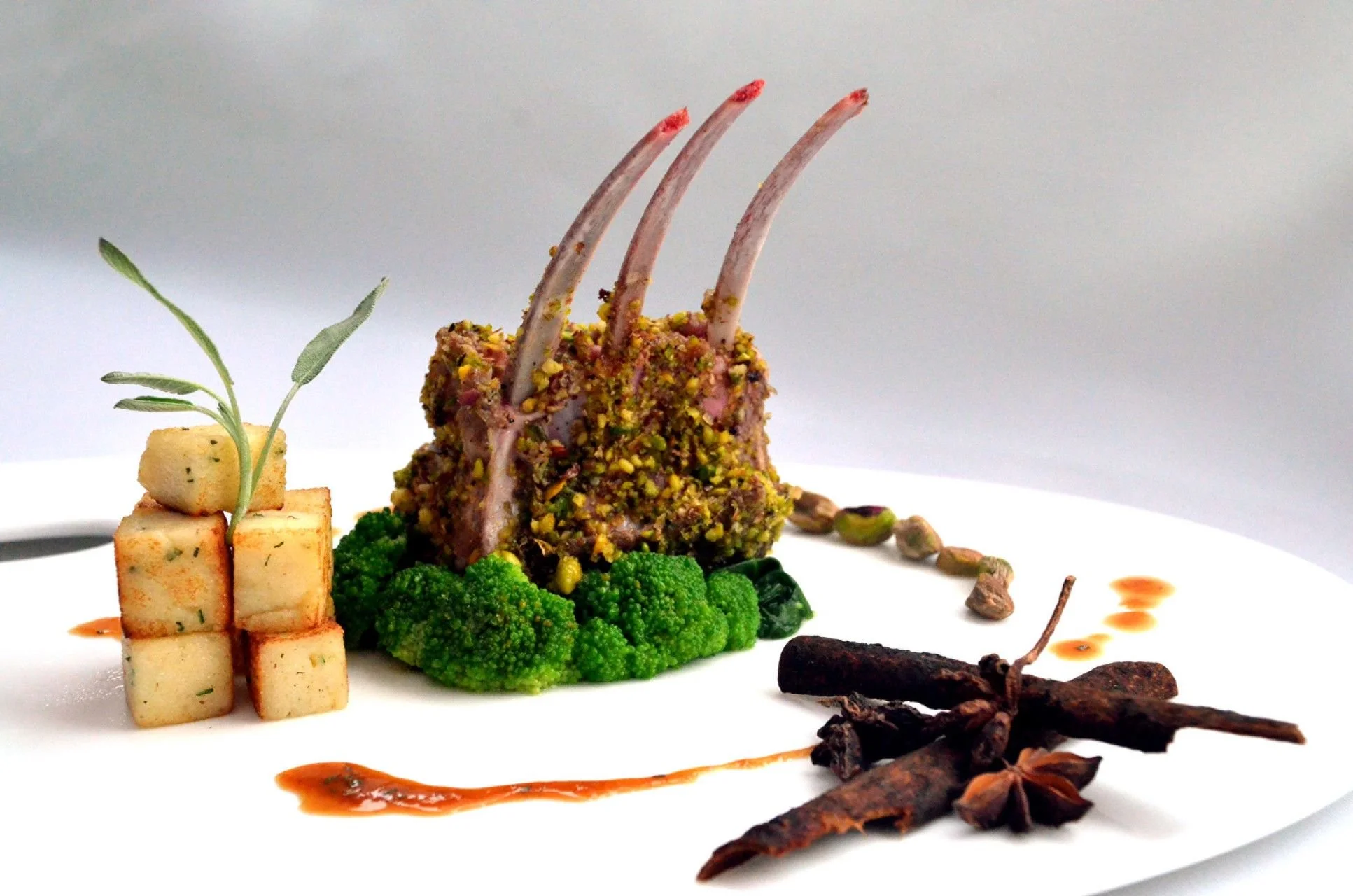 A gourmet dish featuring a breaded piece of meat or fish with three lamb chops on top, garnished with crushed pistachios. The plate includes steamed broccoli, cubed tofu with herbs, and a sprig of sage, with decorative sauces and dried chili peppers.