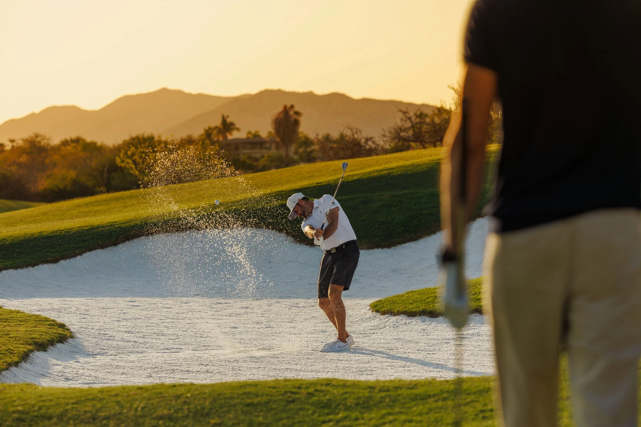A man playing golf sand trap shot during sunset, with another person partially visible in the foreground on a golf course with mountains in the background.