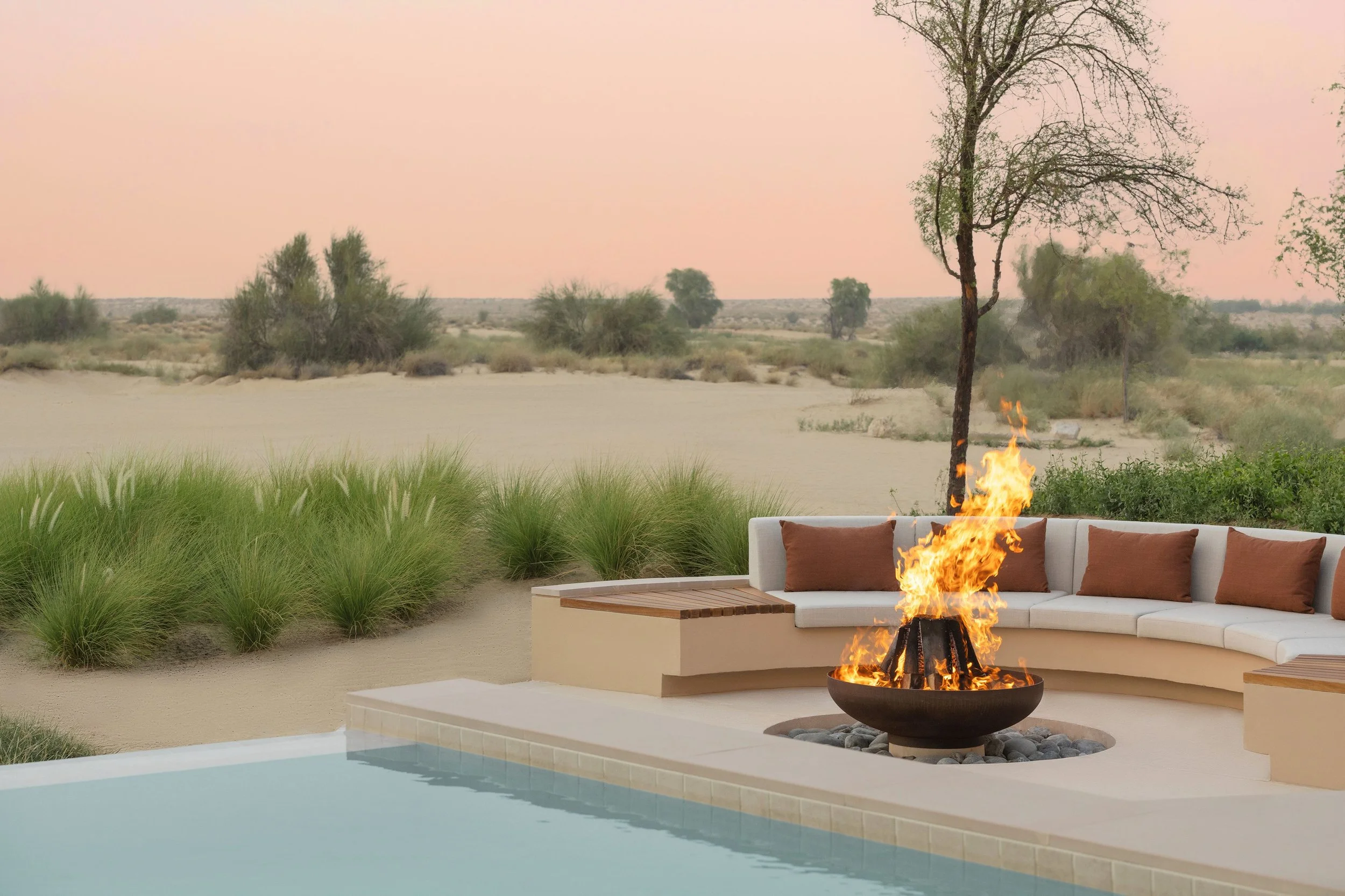 Outdoor patio scene with a swimming pool, circle fire pit with flames, white sectional sofa with brown pillows, desert landscape with shrubs and trees, and a pink sky in the background.