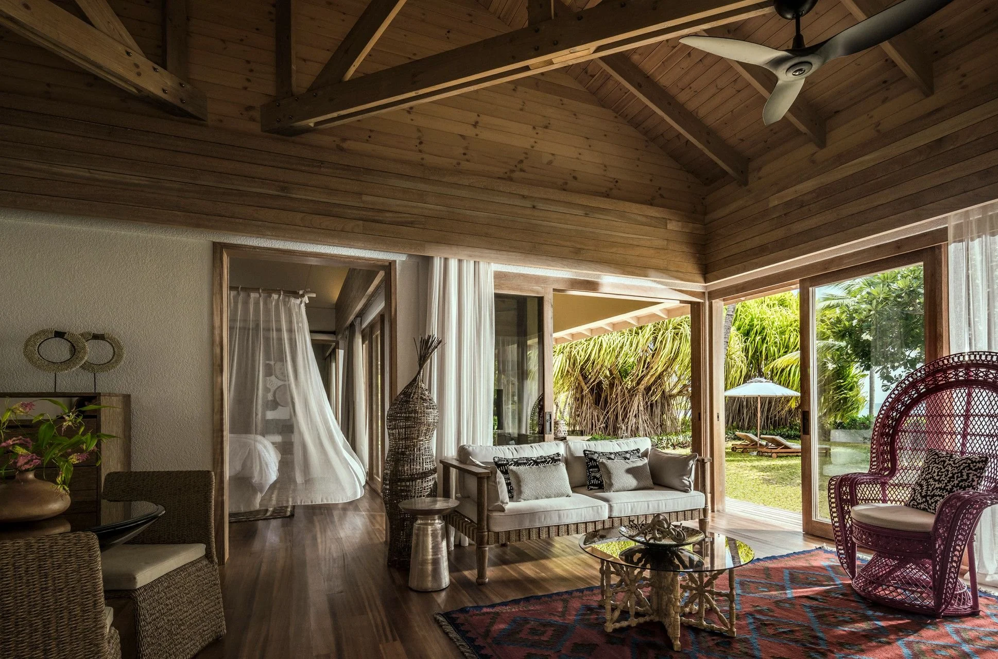 Living room with wooden ceiling, large windows showing outdoor greenery, white sofa, wicker chair, colorful rug, and decorative items.