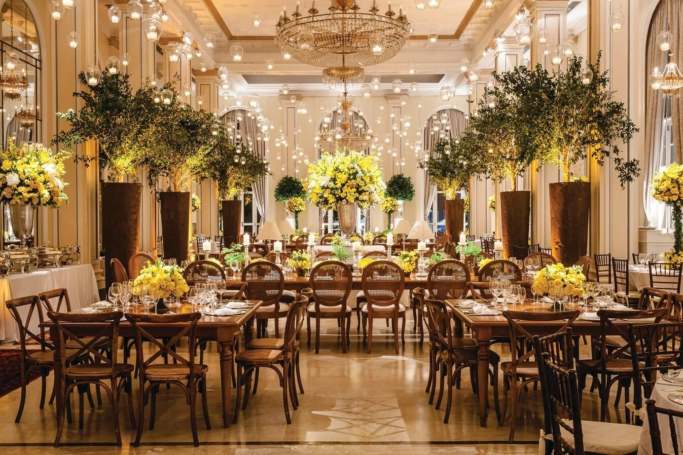 Elegant banquet hall decorated with large potted trees, bright yellow floral centerpieces, and warm lighting with chandeliers and numerous hanging glass lanterns.
