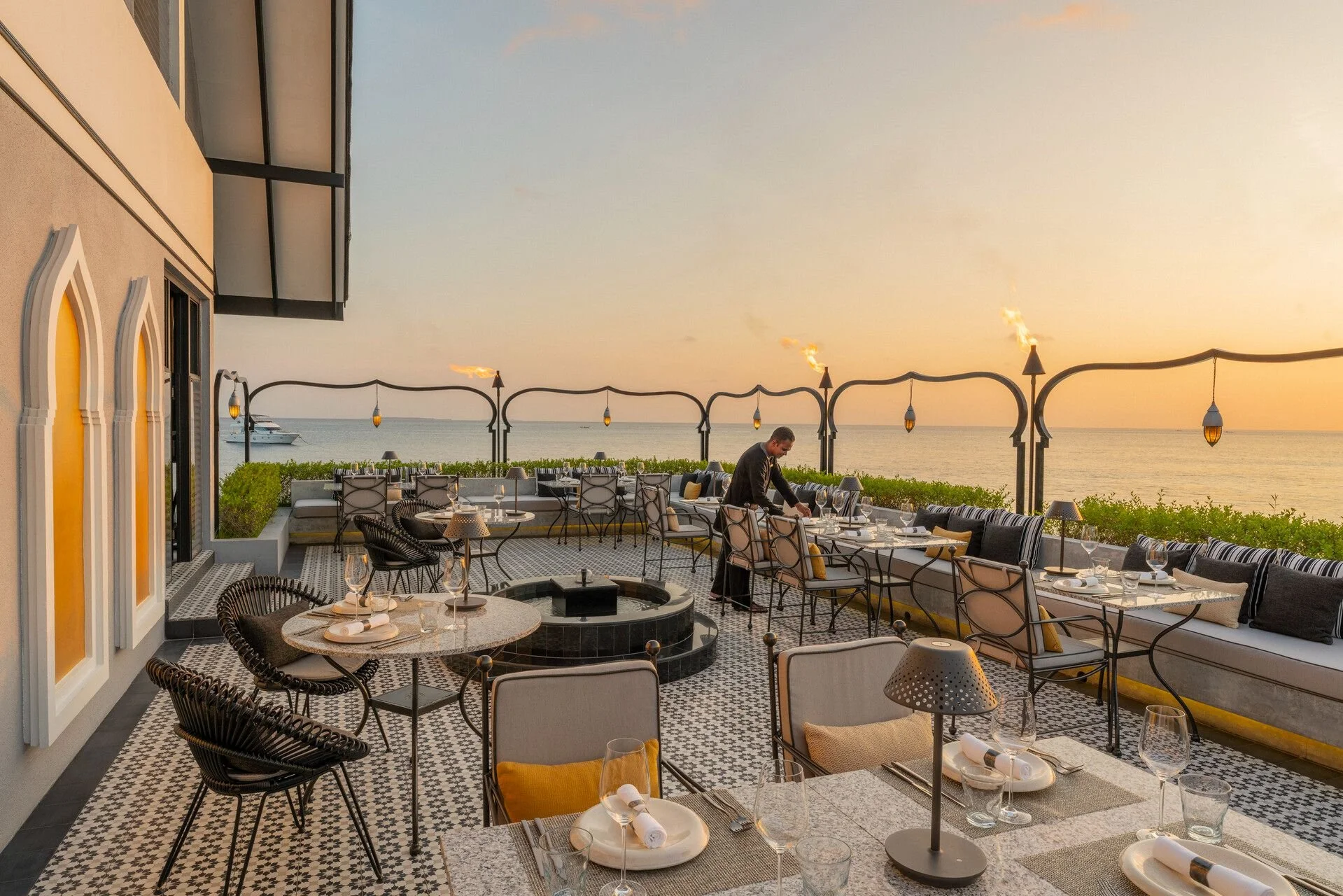 Outdoor restaurant patio overlooking the ocean at sunset, with tables set for dining and a waiter preparing the table, decorative lamps hanging on arching structures, and a yacht on the water in the background.