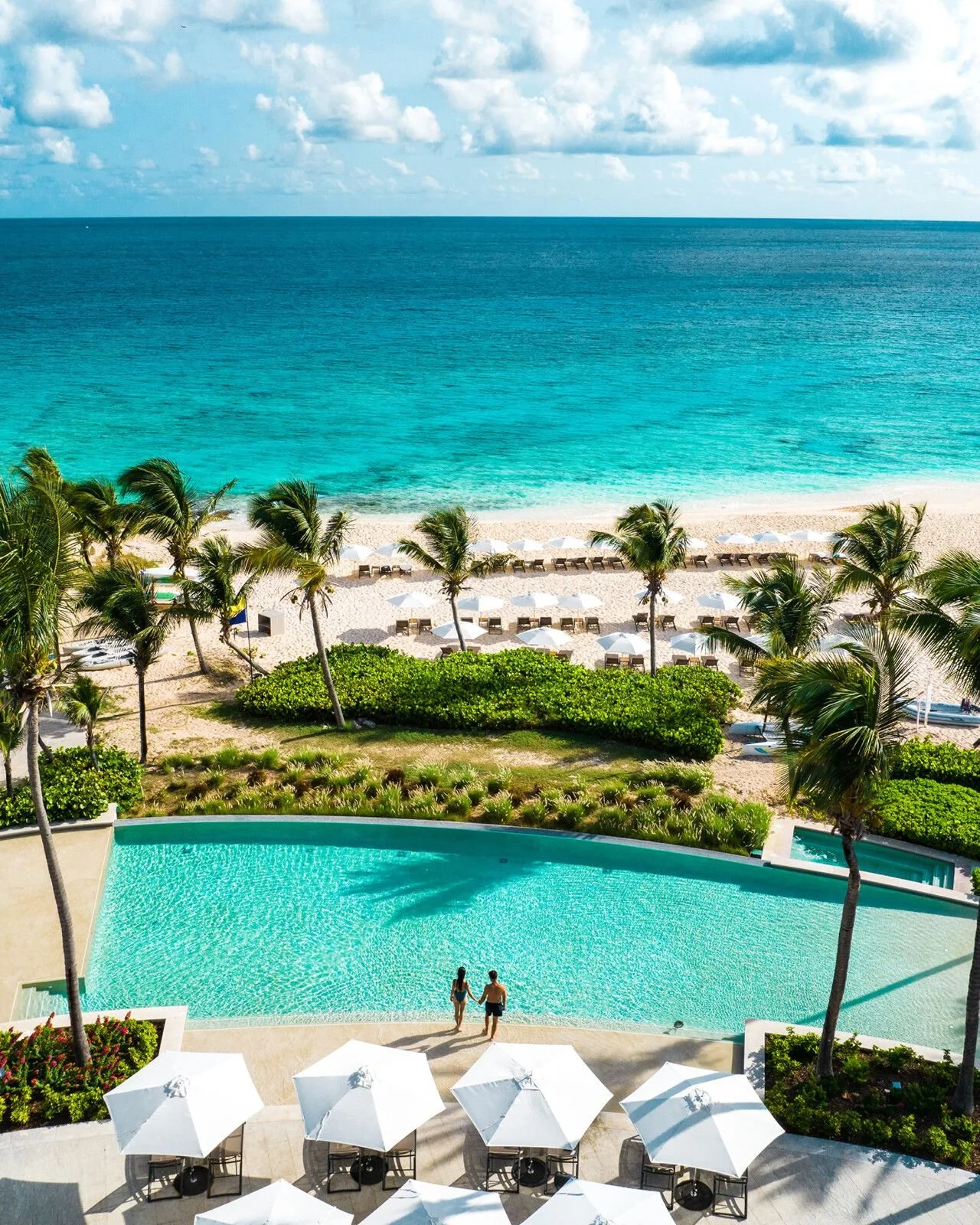 A view from a balcony overlooking a swimming pool, green bushes, and a sandy beach with lounge chairs and umbrellas, beyond the ocean with a partly cloudy sky.