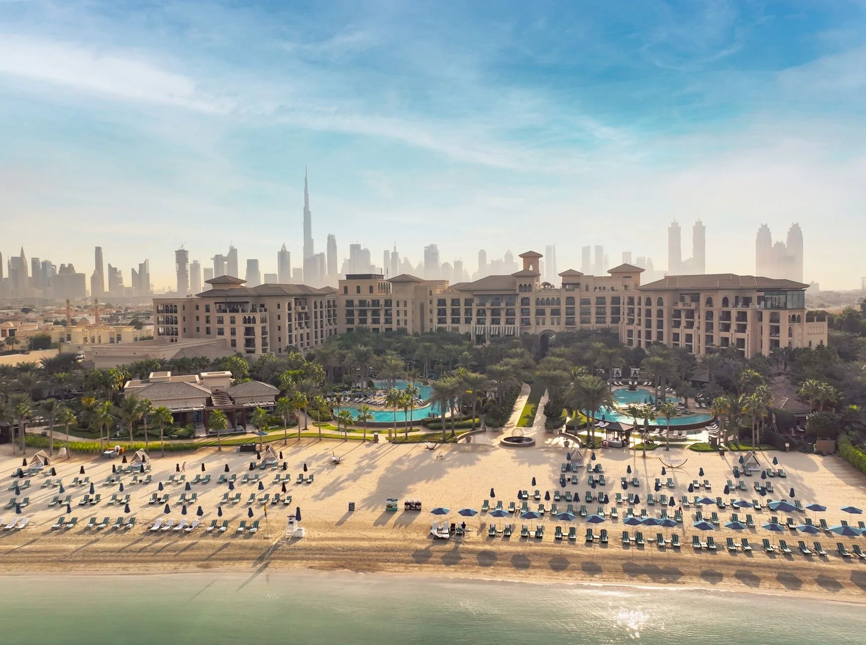 Aerial view of a beachfront resort with pool areas, palm trees, and a city skyline with tall buildings and the Burj Khalifa in the background.