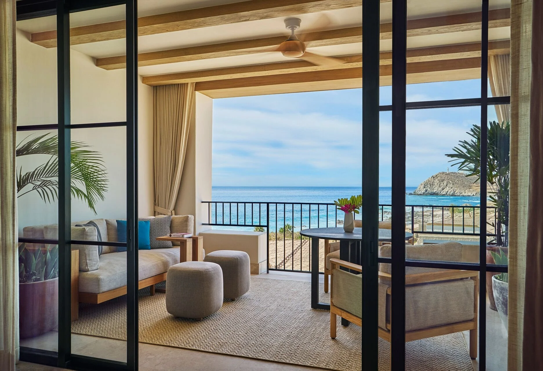 Living room with large glass doors opening to a balcony with ocean view, featuring a sofa, ottoman, chairs, a round table, potted plants, and decorative pillows.