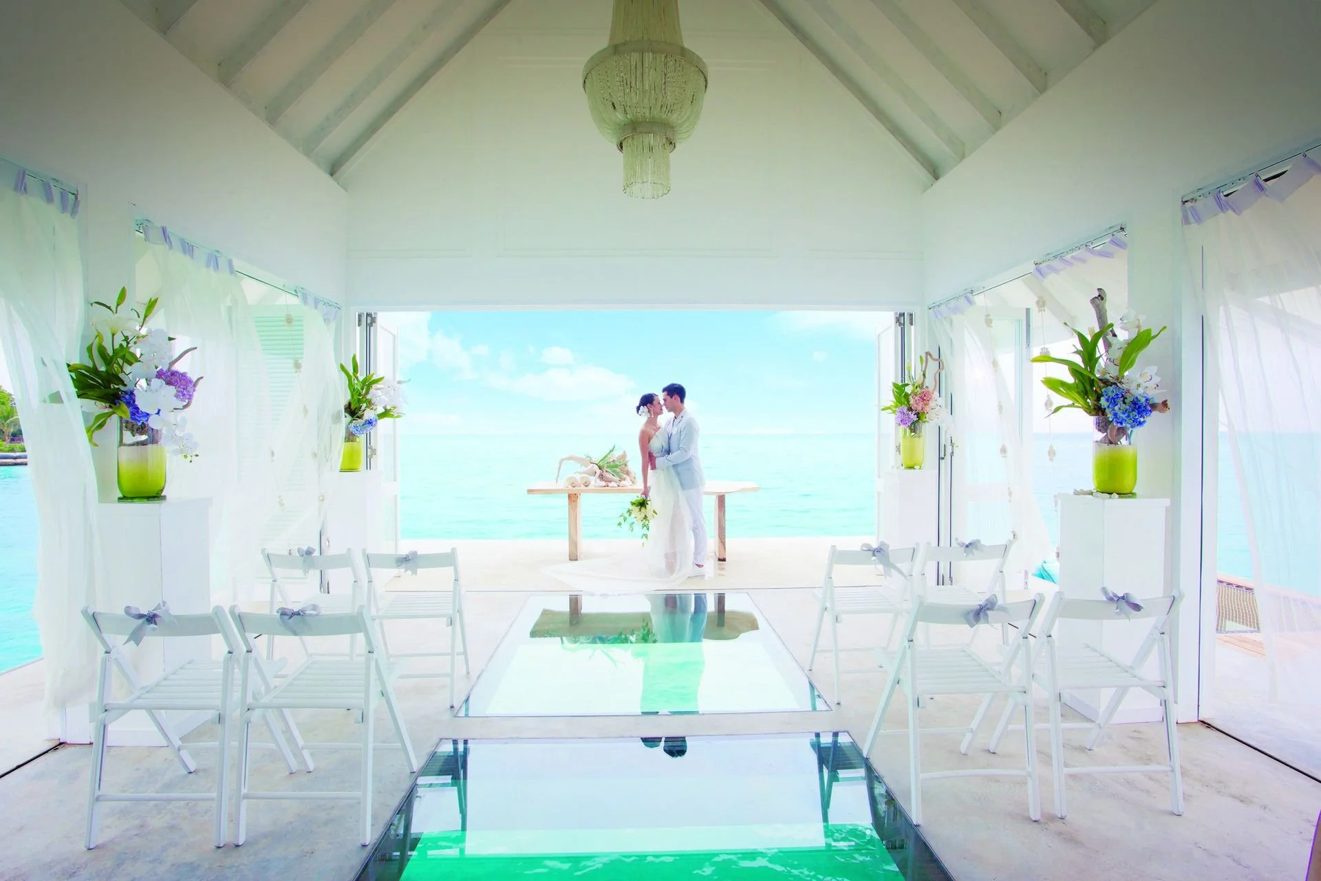 A wedding ceremony setup at a beachside pavilion with white decorations, floral arrangements in green vases, and a bride and groom standing together at the altar with the ocean in the background.