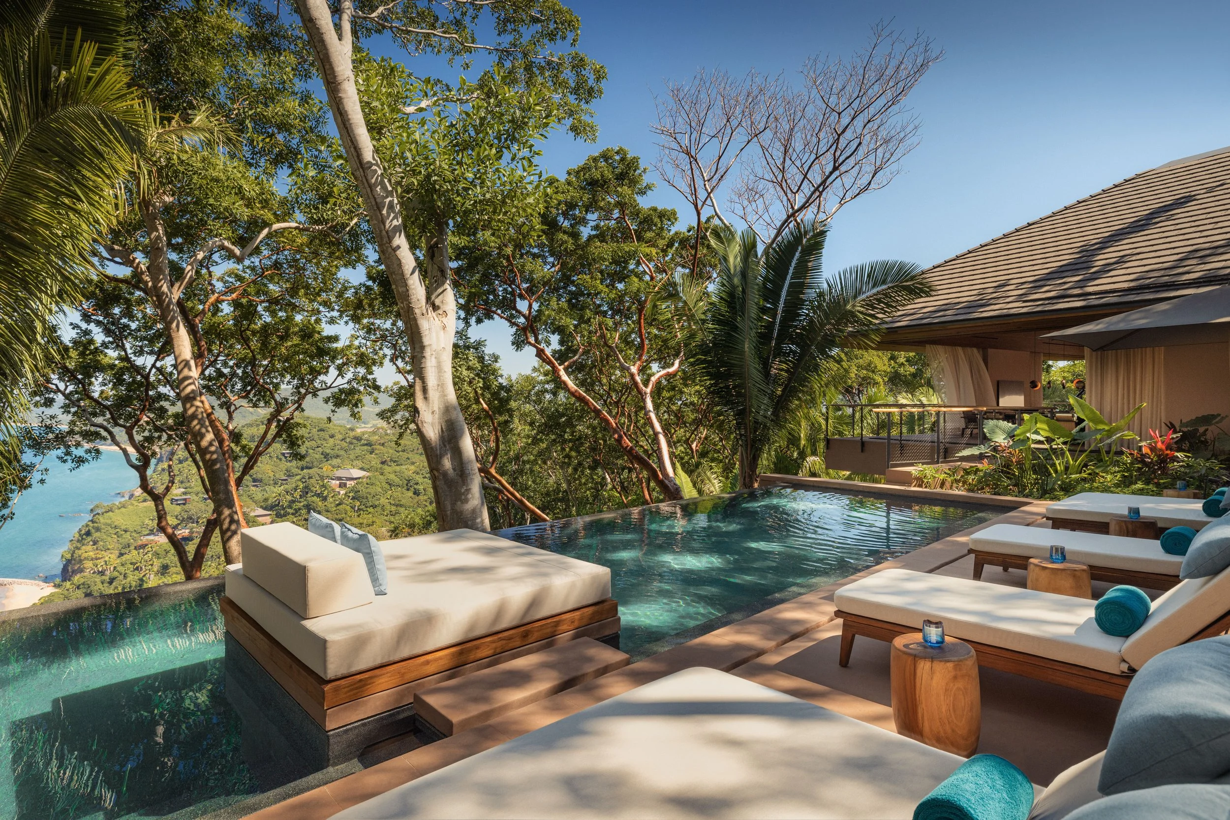 Luxury outdoor pool area with lounge chairs, lush green trees, and a view of a river or ocean in the distance on a clear day.