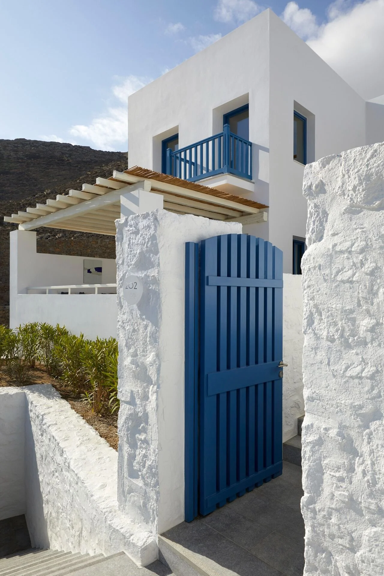 White building with blue accents, a balcony, and a blue gate, surrounded by white walls and a small garden with greenery.