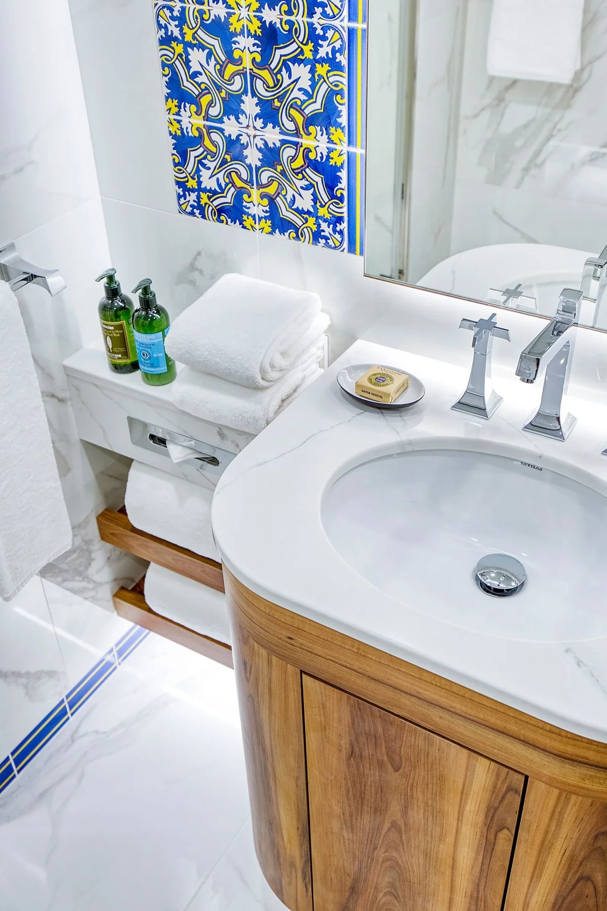A bathroom vanity with a white marble countertop, a carved wooden cabinet, and a round white sink with a chrome faucet. There are two green soap dispensers, neatly folded white towels, a bar of soap, and a mirror with a decorative tiled wall behind it.