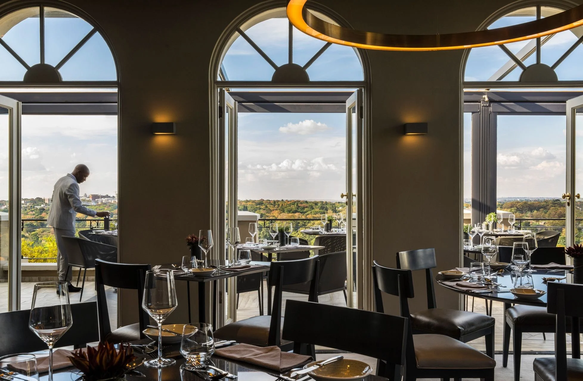 A restaurant with large open windows and a view of the outdoors, set with wine glasses, plates, and silverware. A chef in a white uniform is preparing food inside.