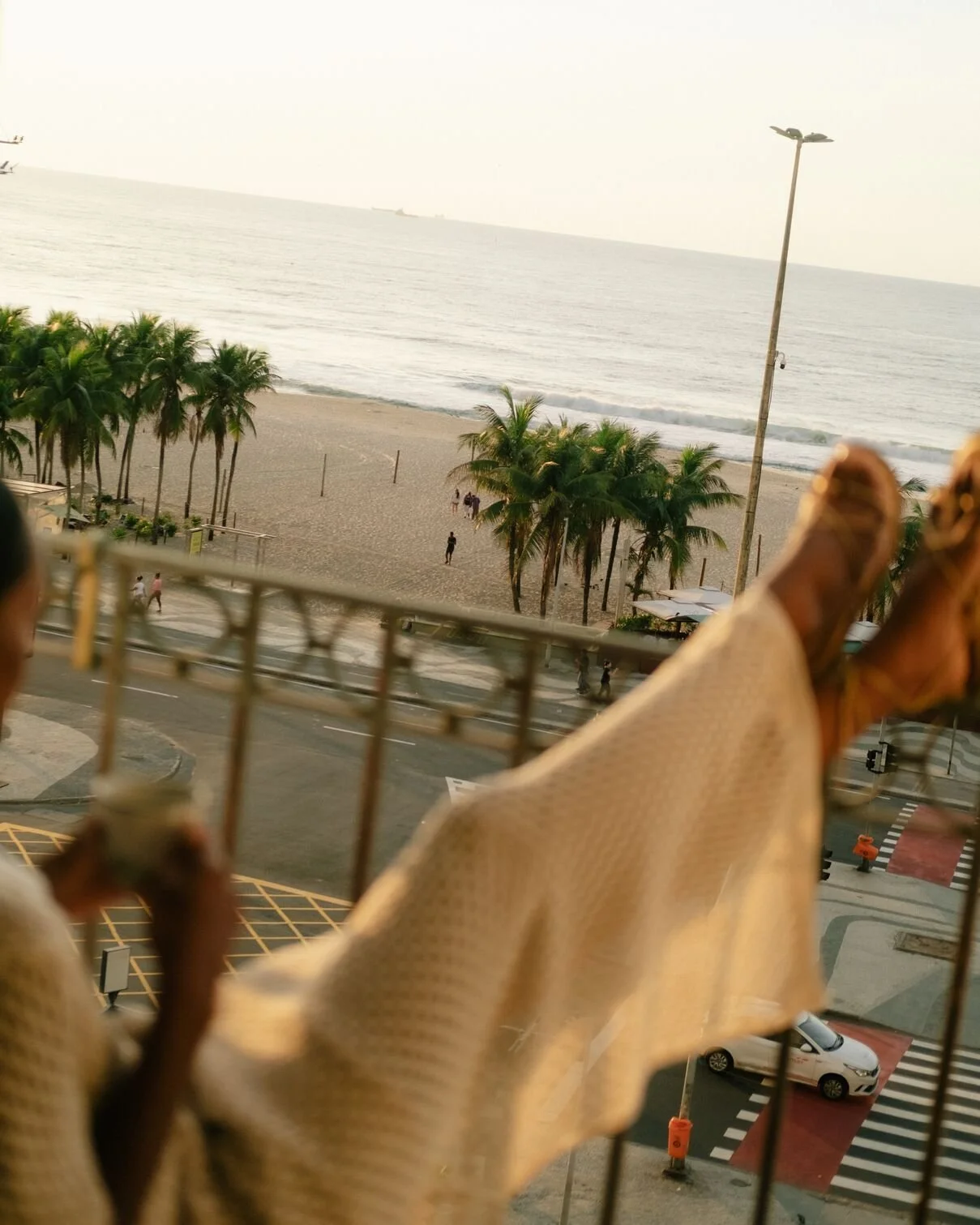 A beach scene with palm trees, shoreline, and ocean in the background, viewed from a balcony with feet in the foreground and part of a metal railing.